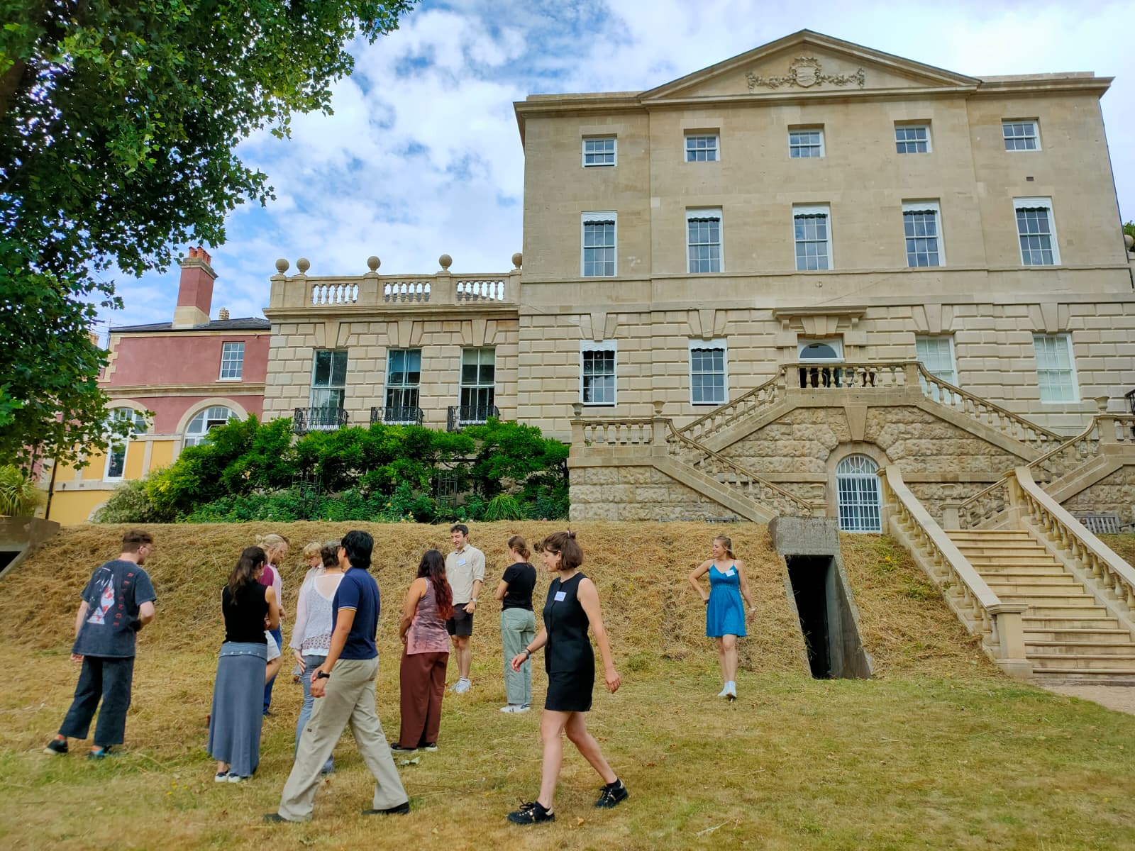 a group of varied students stand outside on some grass, talking animatedly to one another. The students are partaking an activity, stood in a circle.